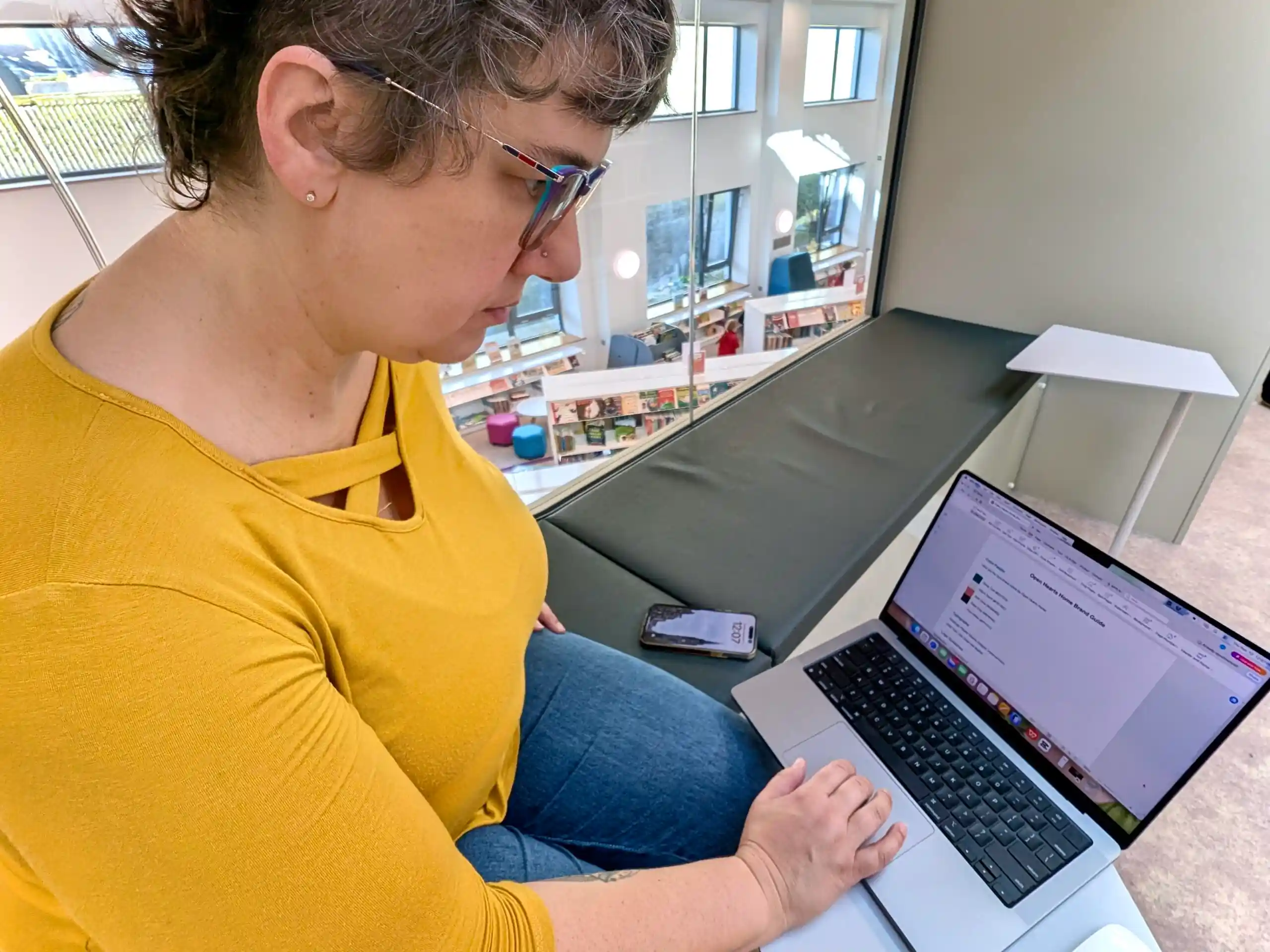 Lisa working on a laptop in a bright library space, reviewing a client brand guide for Nuts and Bolts Marketing.