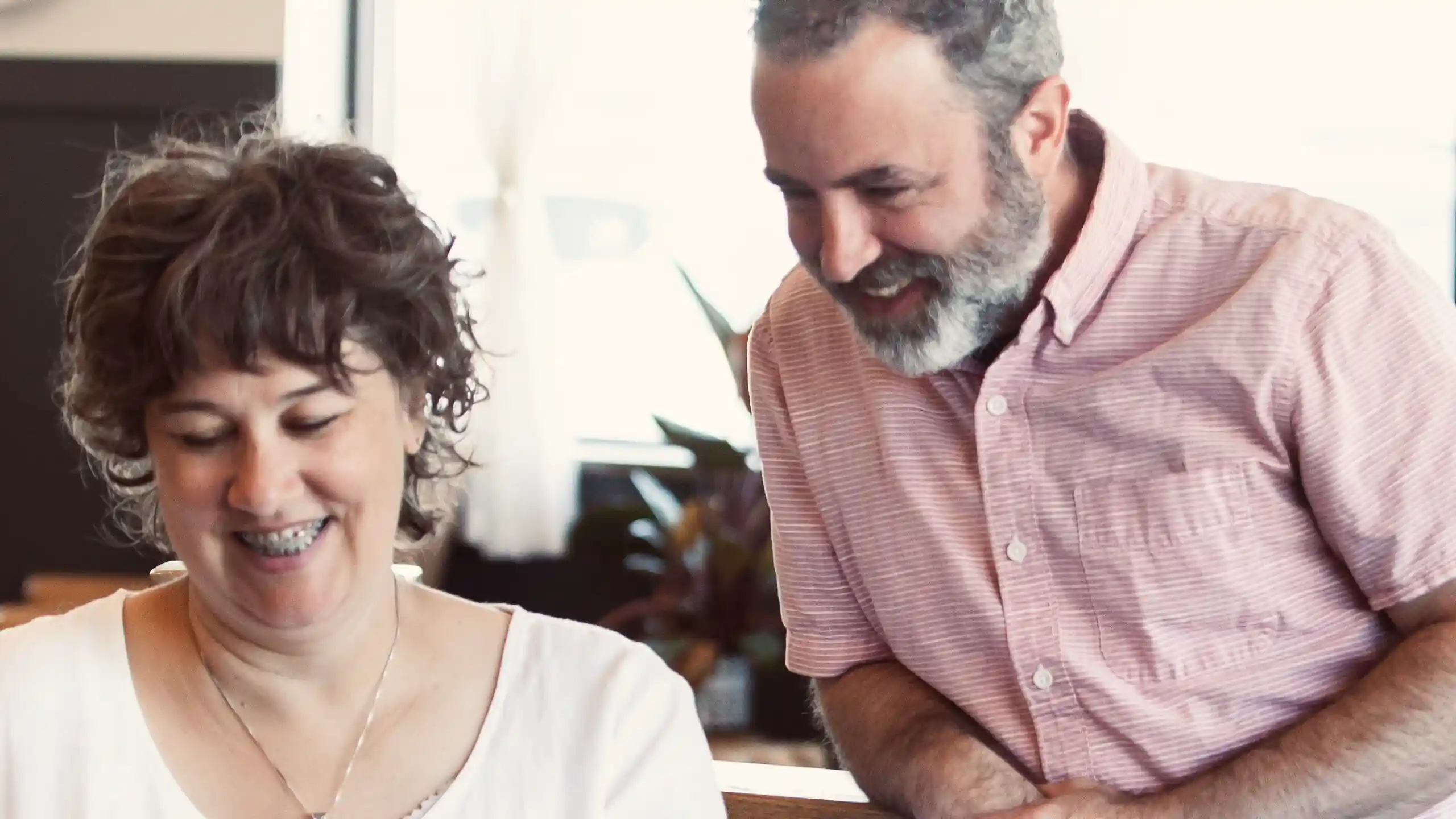 Lisa and Chad smiling and looking at a laptop together inside a bright, cozy café, creating a warm and collaborative moment.