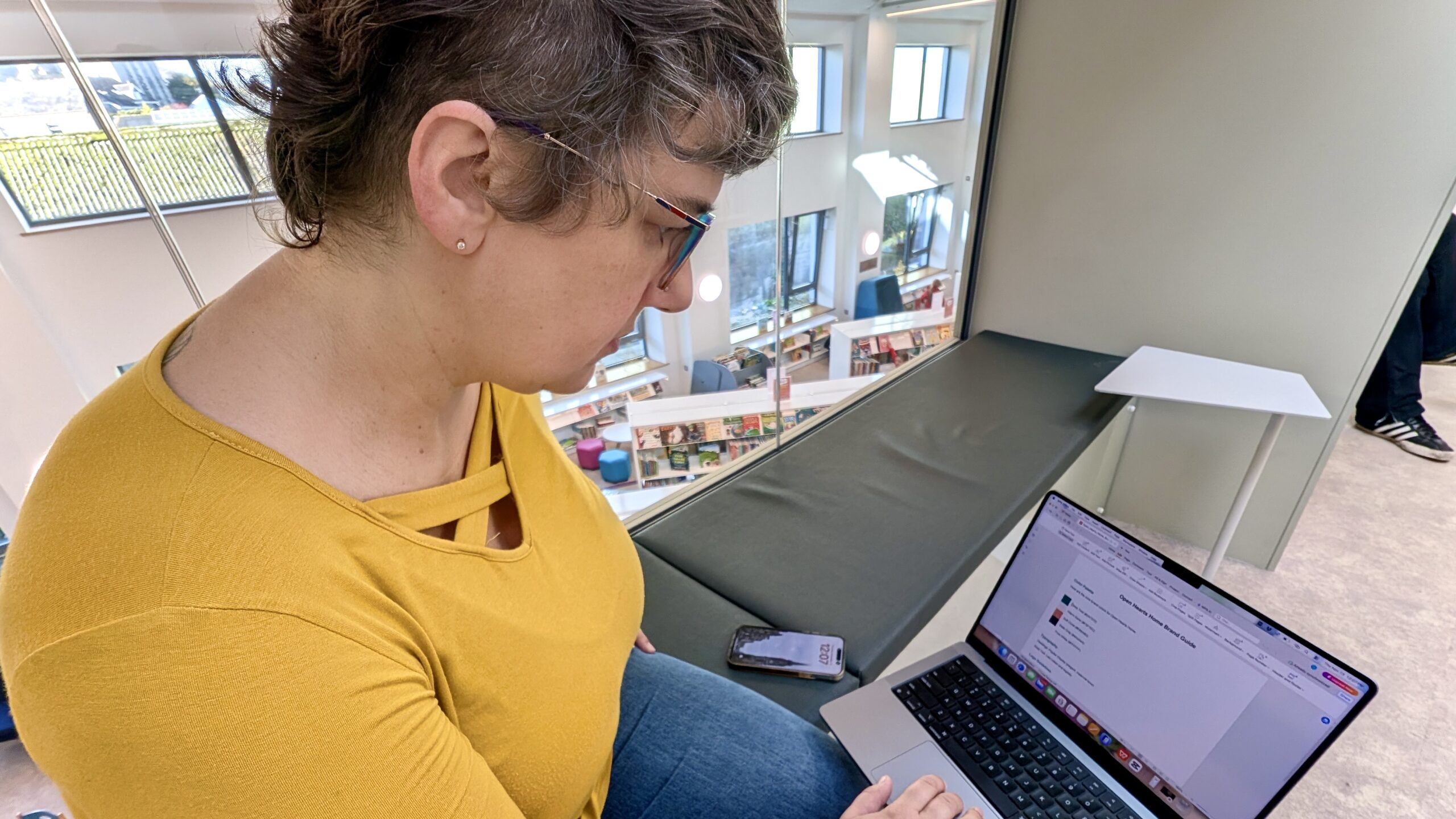 Lisa working on a laptop in a bright library space, reviewing a client brand guide for Nuts and Bolts Marketing.